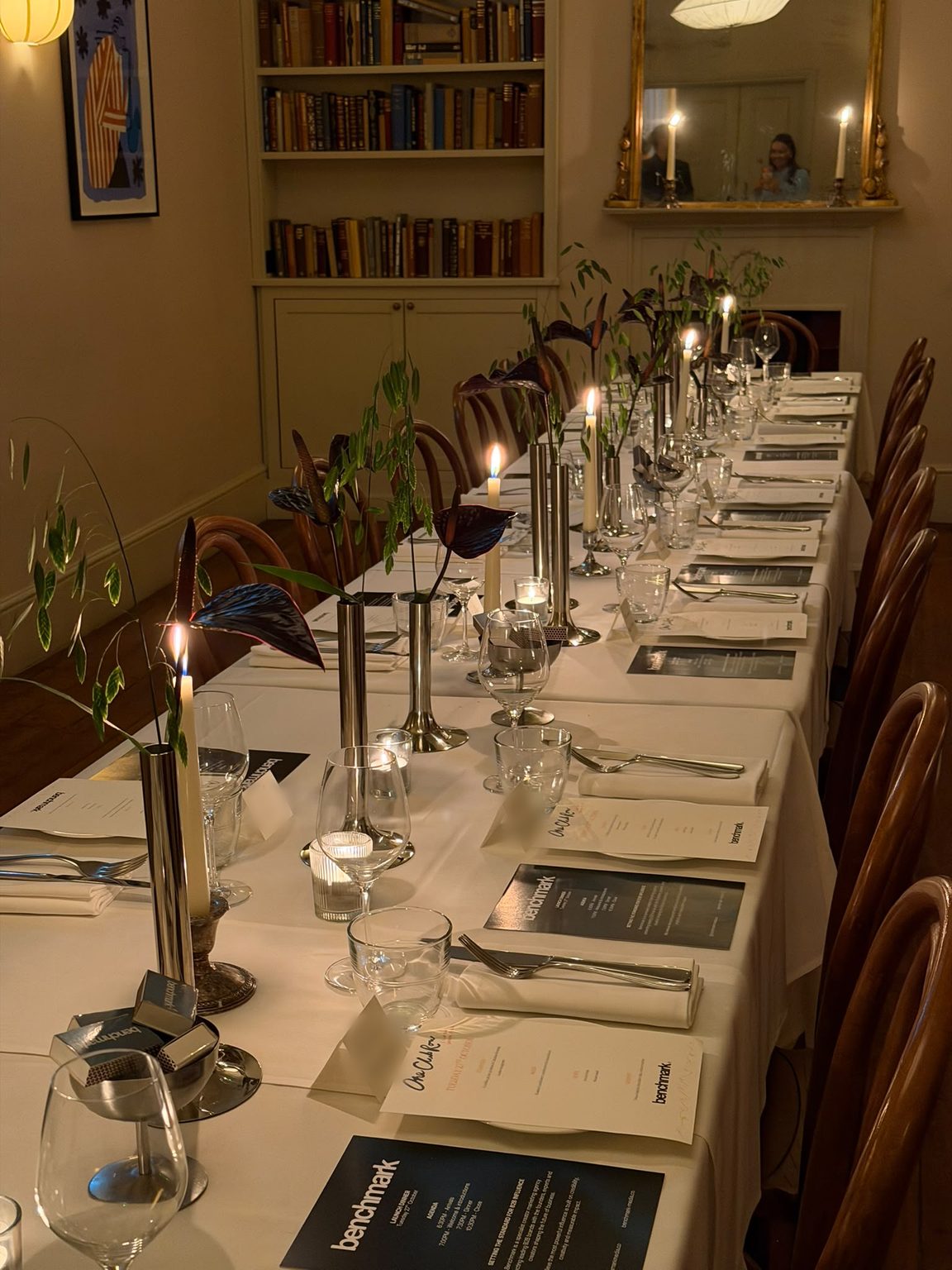 A long candlelit dinner table set with Benchmark menus, silver vases and dark anthuriums at One Club Row